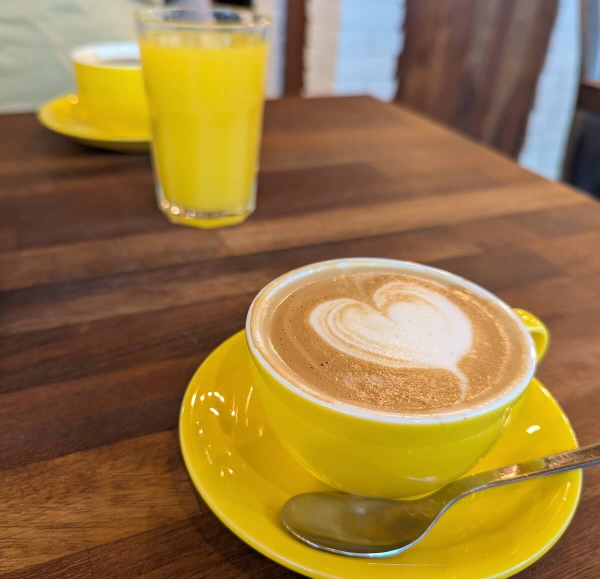 Yellow coffee cups, yellow saucers, orange juice and a silver teaspoon on a wooden table.